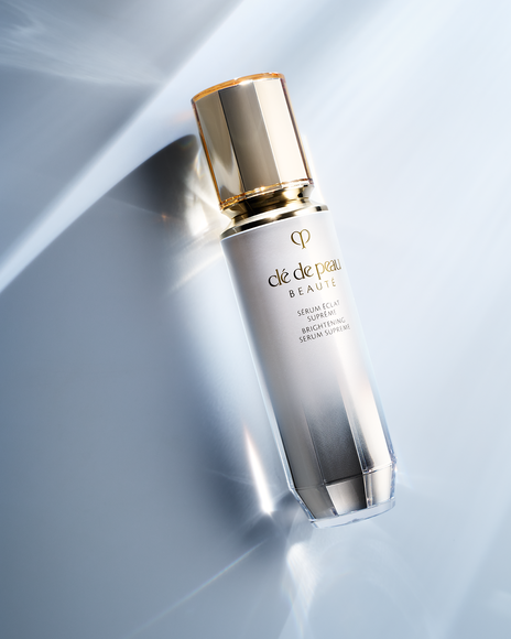 Photo of a brightening serum bottle with a cylindrical shape and multi-faceted design at the base and cap, bathed in diffused light that creates soft metallic reflections on a light grey surface.