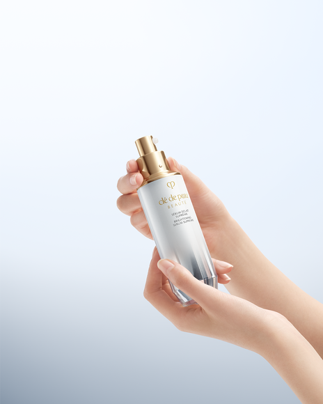 Photo of hands holding a brightening serum bottle with a gold pump, index finger poised to press, lit by soft diffused light against a pale blue gradient.