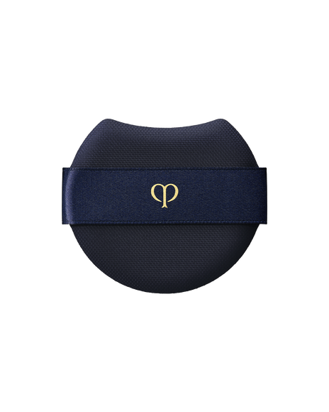 Photo of a "PUFF (RADIANT CUSHION FOUNDATION)" featuring a rounded blue-black makeup puff with a wide, dark blue ribbon across the front and the brand-logo printed in gold at the center.