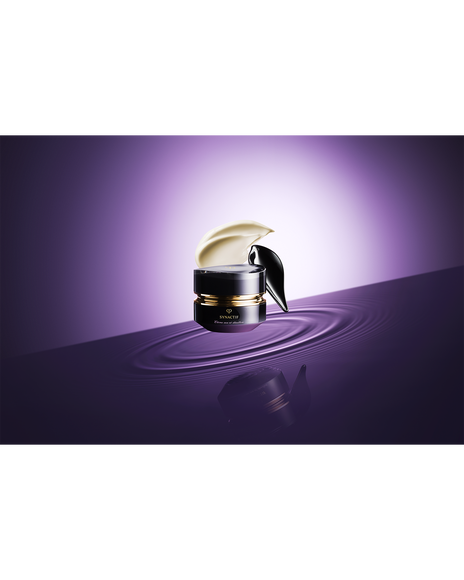 Photo of the black-themed "SYNCACTIF NECK & D&Eacute;COLLET&Eacute; CREAM jar is placed on a purple water surface, showcasing the creamy texture and the appearance of the cream being scooped up with a spatula.