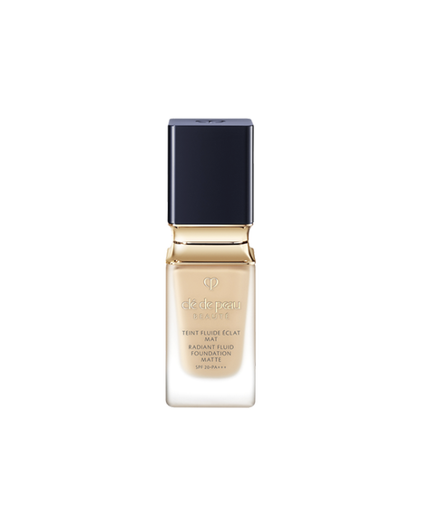 Photo of a bottle of "RADIANT FLUID FOUNDATION MATTE". The liquid foundation comes in a thick, clear glass bottle, featuring a blue-black, square-shaped cap and a gold neck accent. SHADE: O20