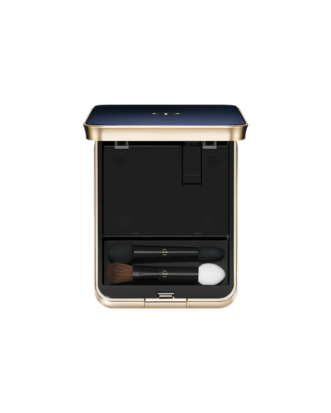 Photo of an exclusive case for "EYE COLOR QUAD" with the lid open. The blue-black lid features a gold trim. Inside, the top half has an empty compartment, while the bottom section holds two makeup applicators: a small brush and sponge-tip applicators.
