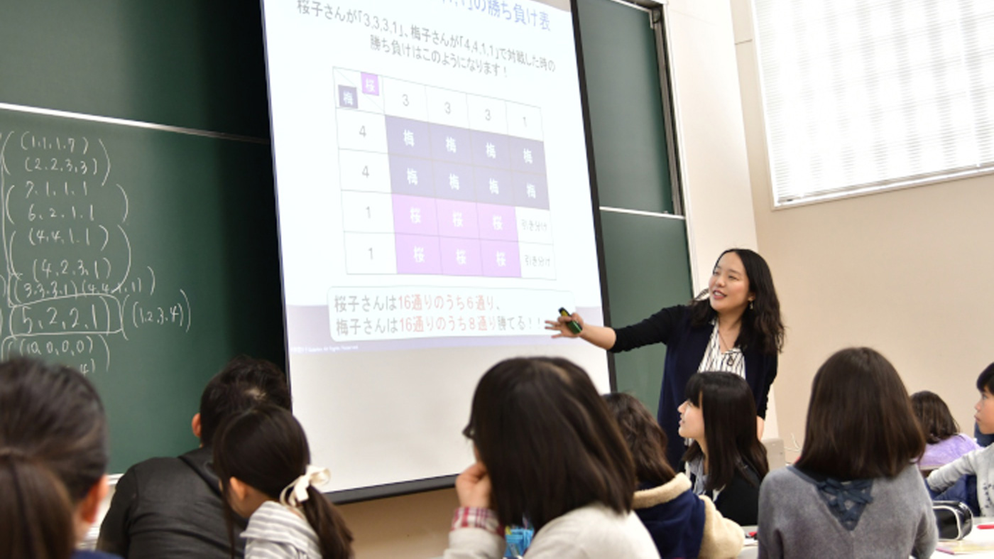 Photo of Sachiko Nakajima teaching a group of students in a classroom, pointing to a projected slide at the front of the room.