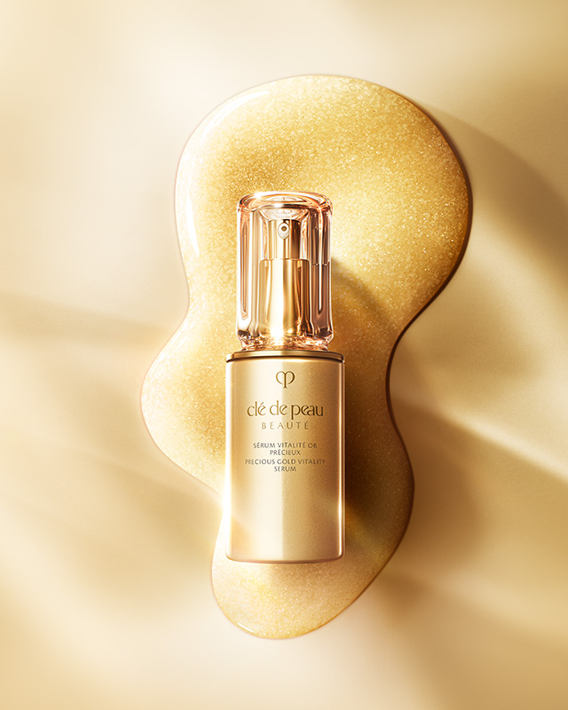 相片顯示Cl&eacute; de Peau Beaut&eacute;珍稀黃金賦活精華的瓶身，優雅置於注入了24K黃金精萃的豐潤閃亮精華上，彰顯其奢華非凡的特質。