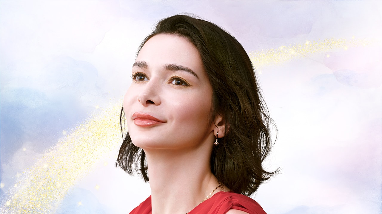 Photo of a model wearing a red top, with yellow and gold eye makeup and red lipstick. Rainbow pattern appears in the background. 