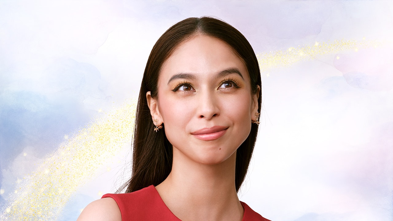 Photo of a model wearing a red top, with brown-toned eye makeup and pink lipstick. Rainbow pattern appears in the background. 